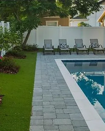 A modern rectangular pool set into a paver deck, with fresh lawn, garden beds and lounge chairs along the far edge.