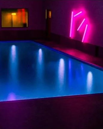 The same style of pool lit blue at night, with pink neon wall lighting reflecting across the water.