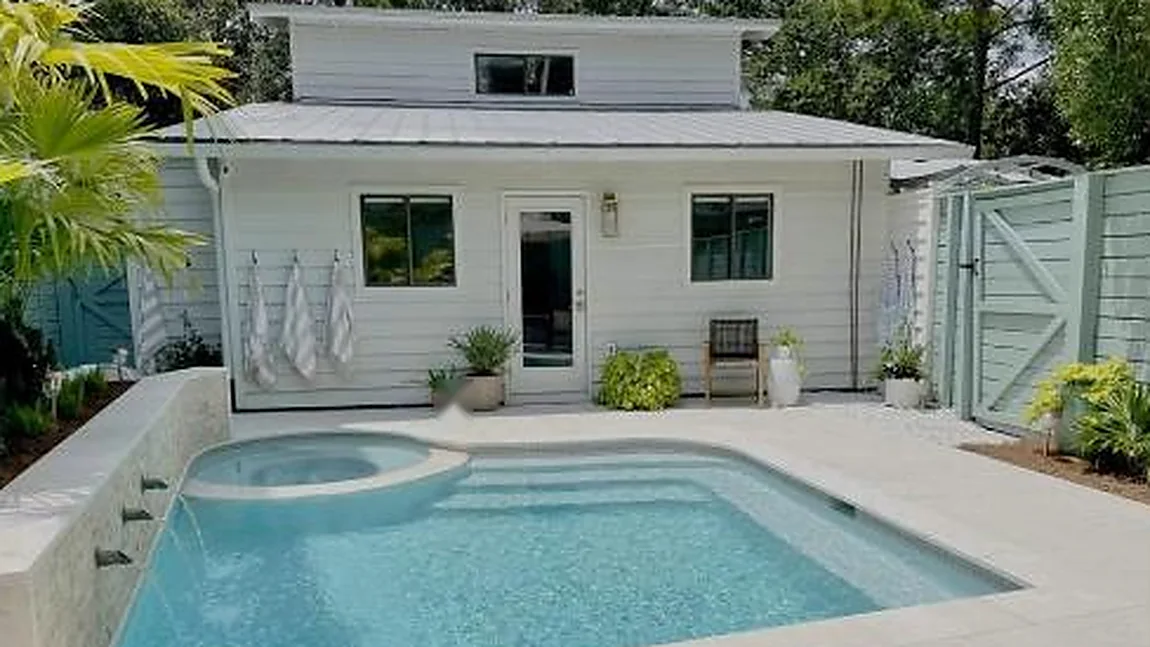 A backyard pool and spa built by Lagoon Pools of South Florida, with water spouts spilling into clear blue water beside a white cottage.